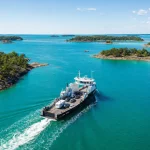 A small car ferry crossing bright water between wooded islands, seen from the air