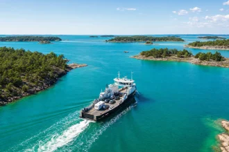 A small car ferry crossing bright water between wooded islands, seen from the air