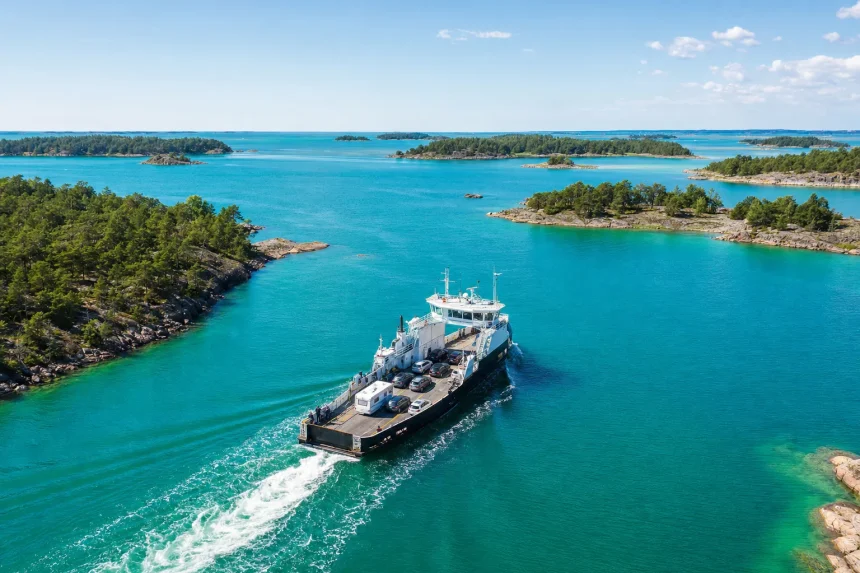 A small car ferry crossing bright water between wooded islands, seen from the air