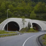 A twin-bore road tunnel portal cut into a green forested hillside in clear morning light