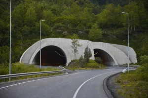 A twin-bore road tunnel portal cut into a green forested hillside in clear morning light
