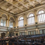Daylight through tall arched windows onto the ornate ceiling of a legislative chamber