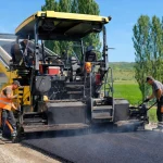 A road crew laying fresh asphalt with a paver and roller on a rural road in summer