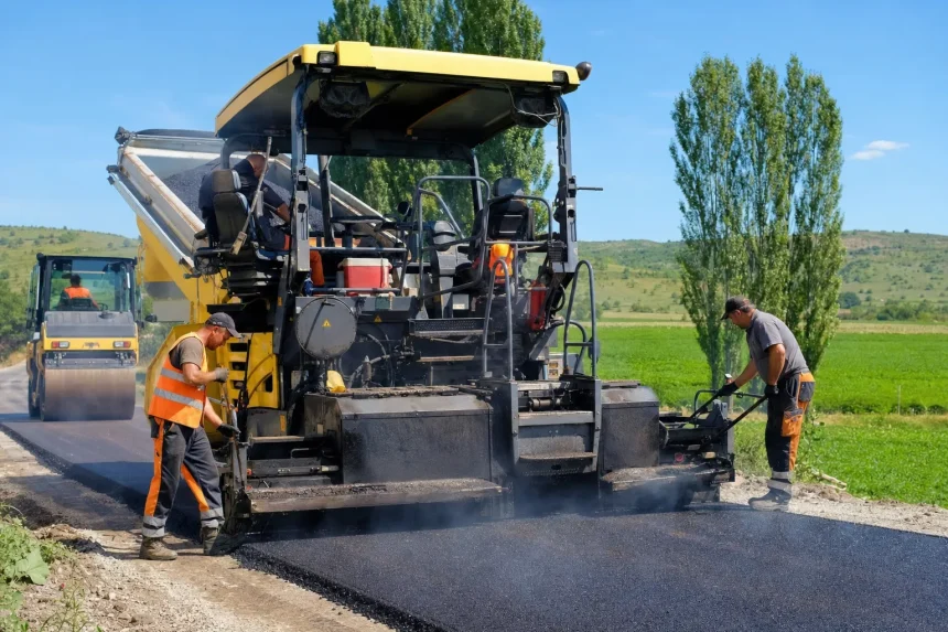 A road crew laying fresh asphalt with a paver and roller on a rural road in summer